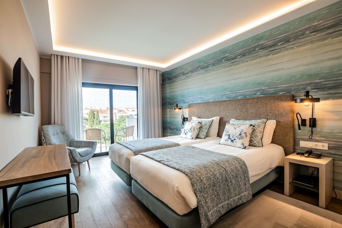 A modern hotel room with two beds, a desk, a chair, and a view of the outdoors.