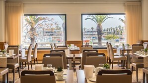 A hotel dining area with tables set for dining, large windows offering views of a marina and palm trees. - Marina Rio (Lagos)