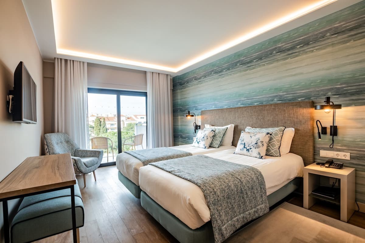 A hotel room with two beds, a desk, a chair, a TV, and a balcony with curtains.