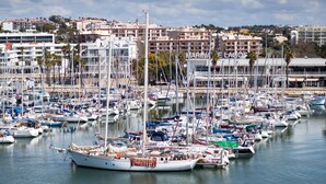 A marina with numerous boats docked, a building labeled "MARINA DE LAGOS" in the background, and a cityscape with residential buildings. - Marina Rio (Lagos)