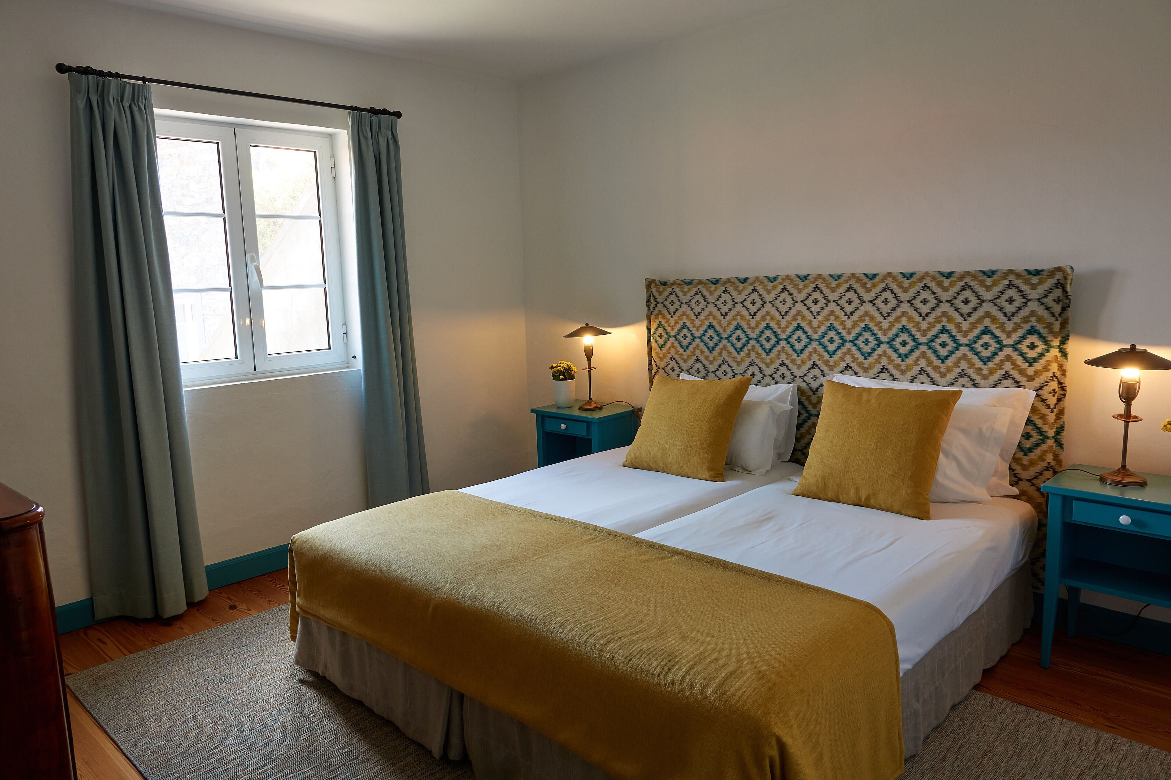 A double bed with a patterned headboard, two bedside tables with lamps, a window with curtains, and a wooden floor.