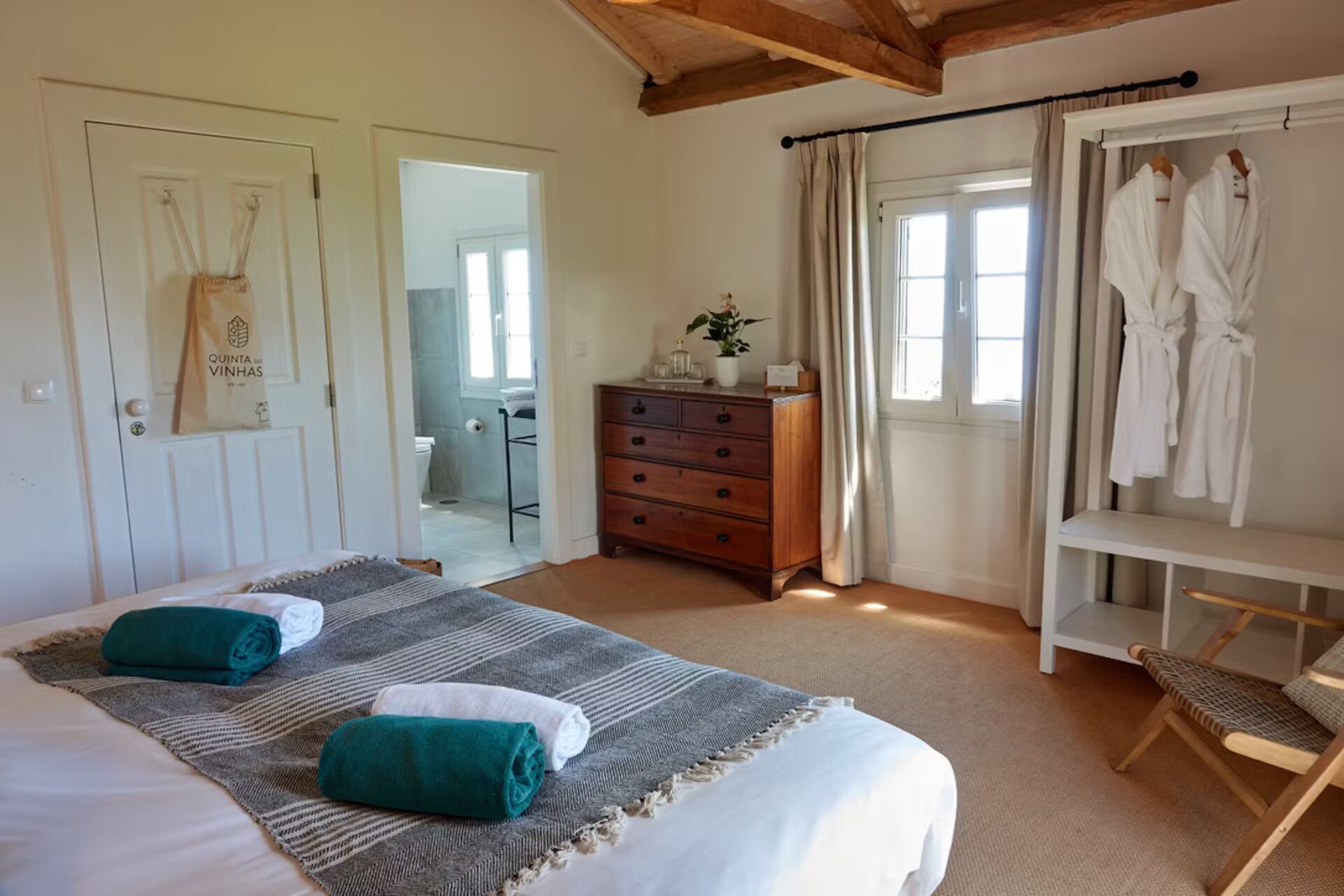 A bedroom with a bed, two rolled towels, a wooden dresser, a bench, and a robe rack.