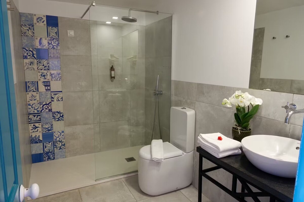 Un baño moderno con ducha envuelta en vidrio, inodoro blanco, lavamanos con un florero y un espejo.