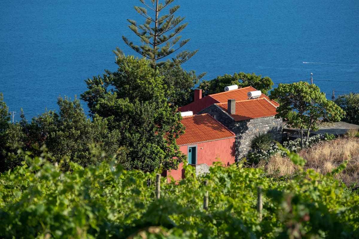 A red building with a stone foundation, surrounded by lush greenery and overlooking a body of water.