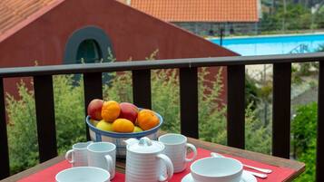 A table set with white cups, bowls, and a bowl of fruit on a balcony with a view of a swimming pool and buildings.