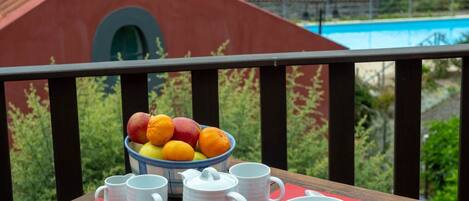 A table set with white cups, bowls, and a bowl of fruit on a balcony with a view of a swimming pool and buildings.