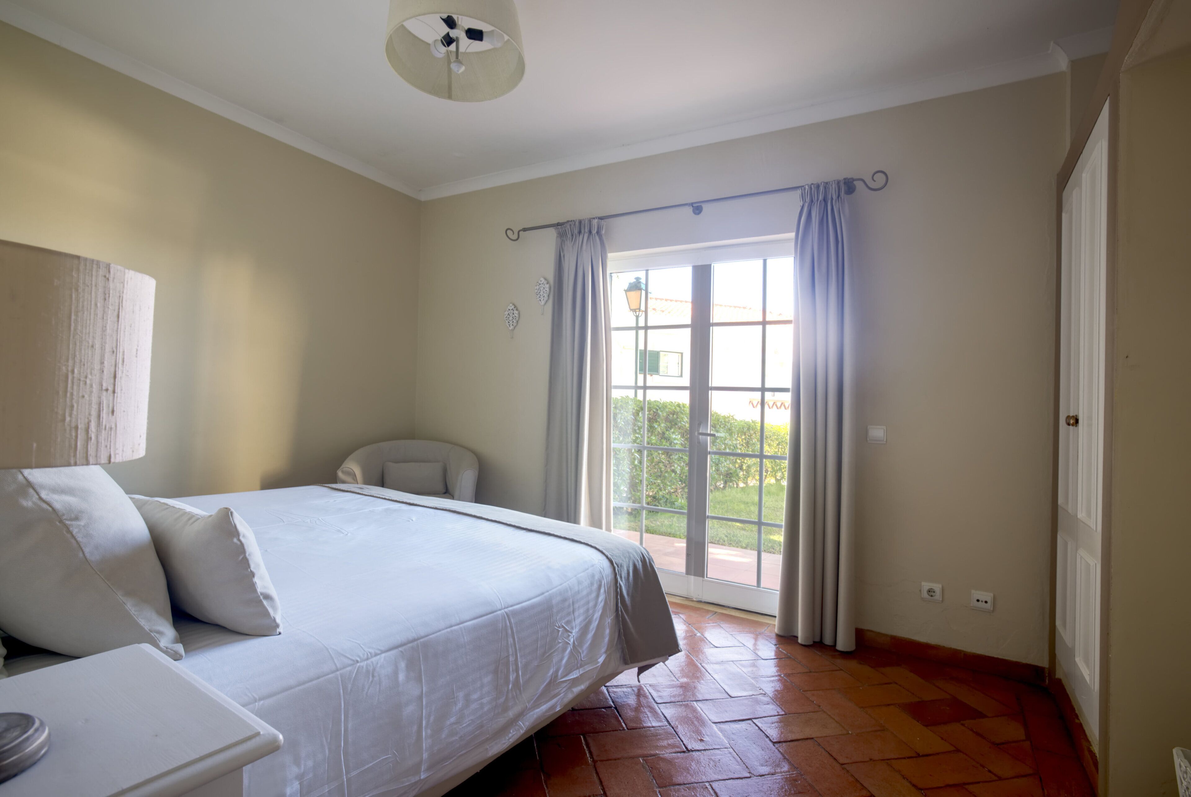 Een slaapkamer met een bed, een stoel, een lamp en een raam met gordijnen.