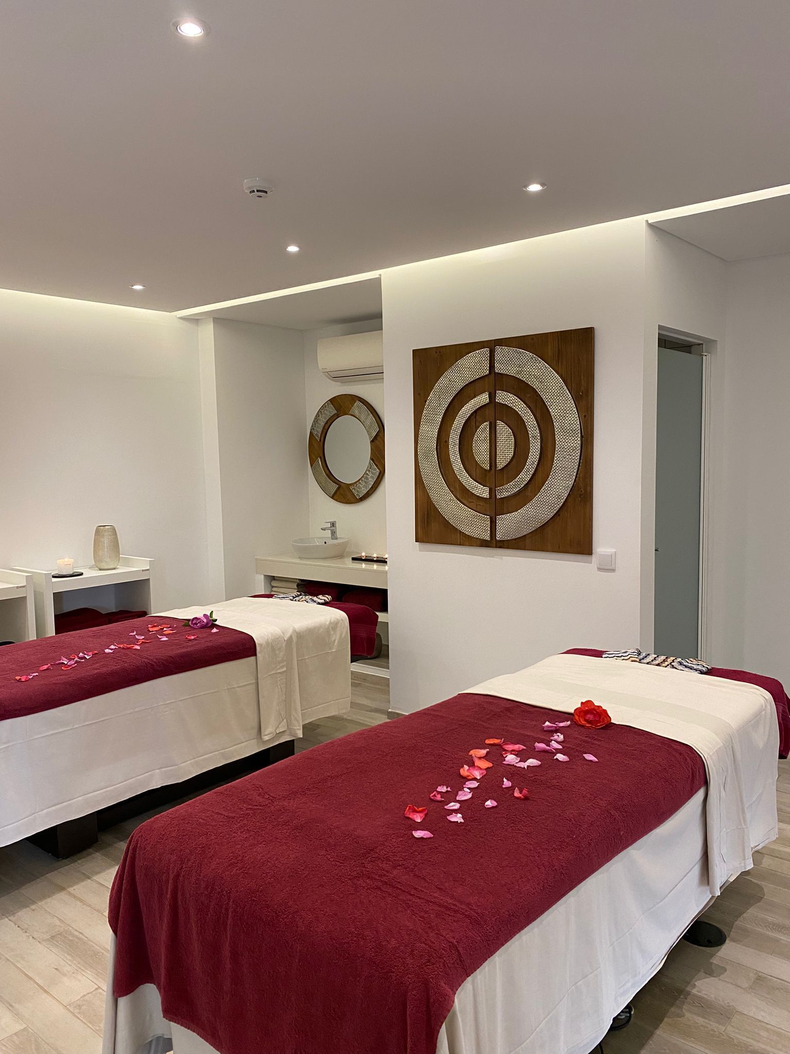 Uma sala de spa com duas camas, cada uma com uma toalha vermelha e pétalas de rosa. Há um espelho, uma pia e um vaso com uma vela sobre uma mesa.