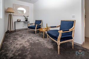 A room with two blue upholstered chairs, a small round table with a bottle and glasses, a floor lamp, and a mirror on the wall.