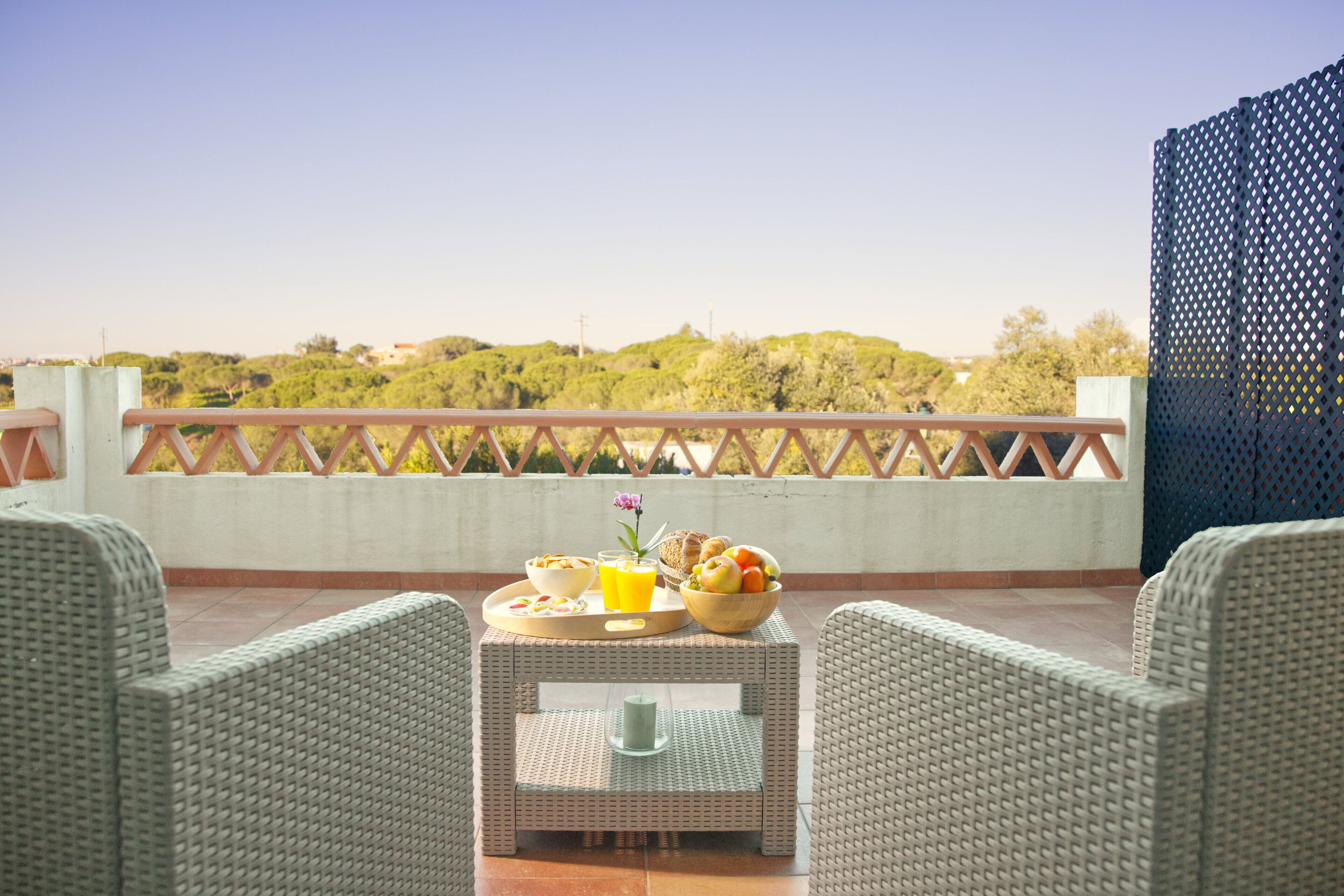A balcony with wicker furniture, a tray with breakfast items, and a view of greenery and buildings in the distance.