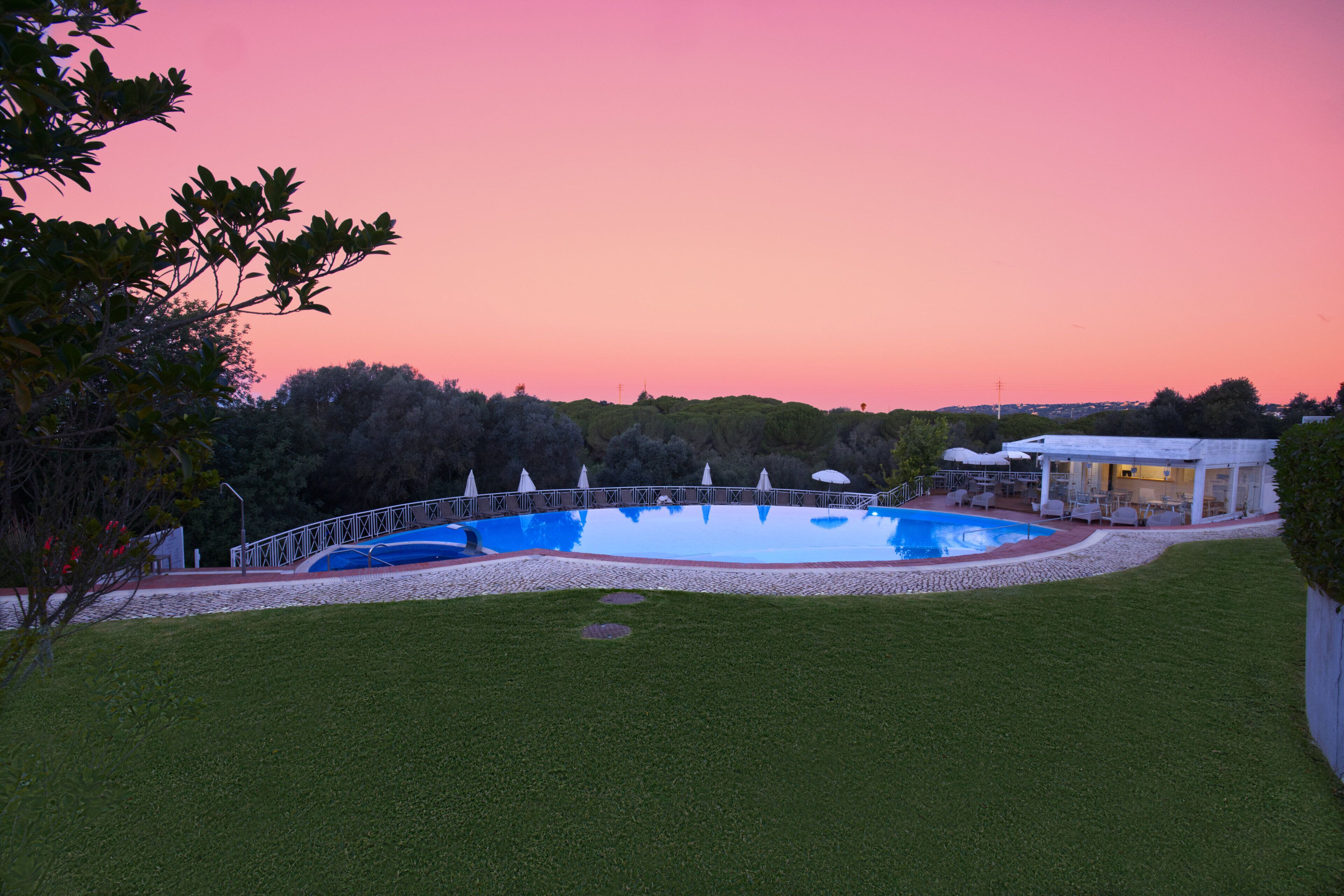 A swimming pool surrounded by a white fence, with a covered patio area and a building in the background.