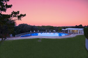 A swimming pool surrounded by a white fence, with a covered patio area and a building in the background.