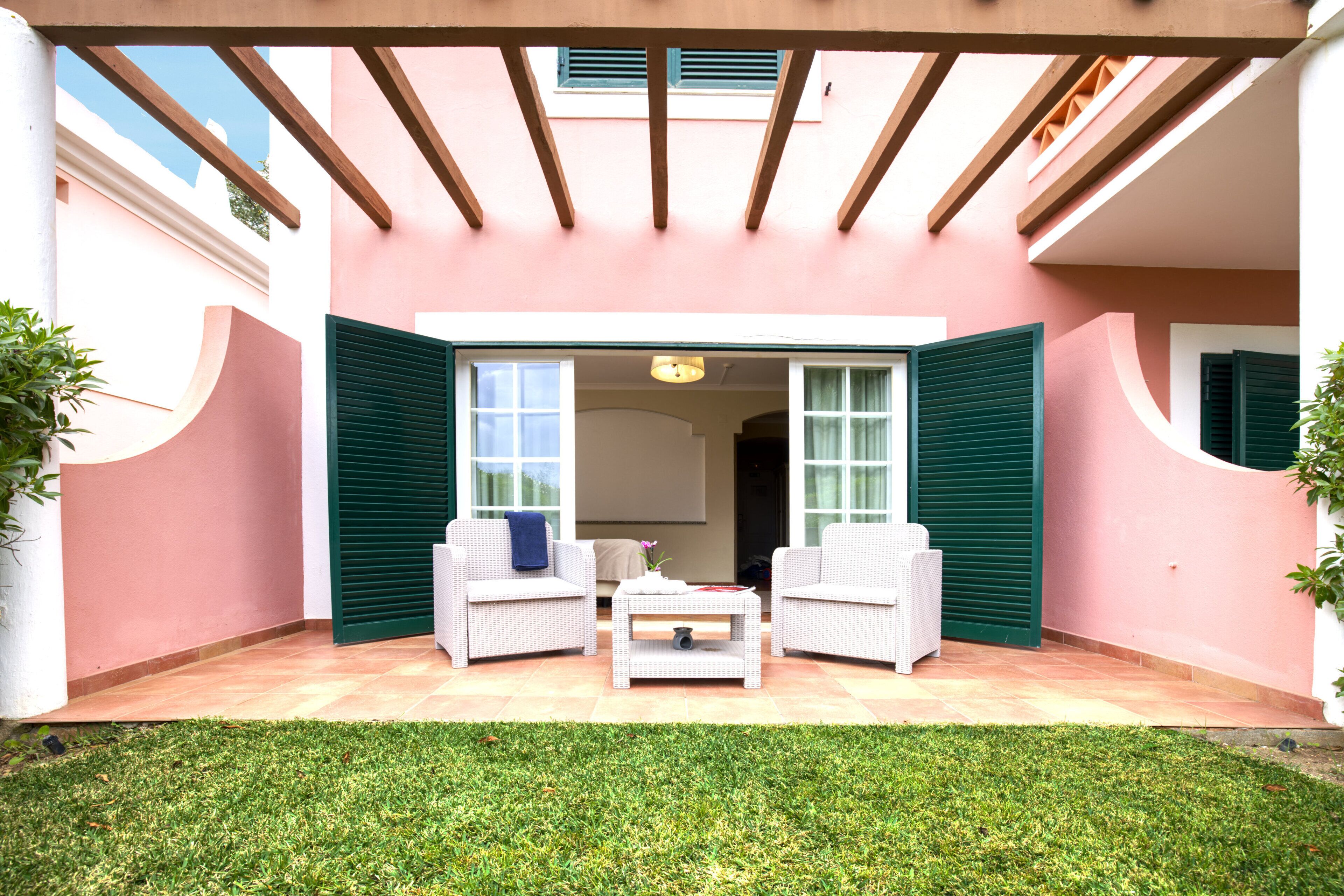 A covered patio area with two white wicker chairs and a small table, surrounded by a pink wall and green shutters.