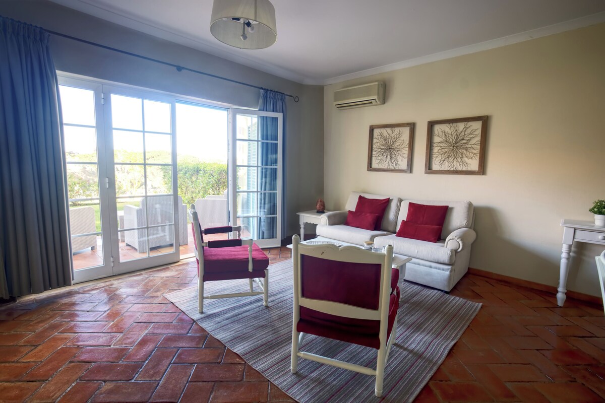 A room with a white sofa, red cushions, a white chair, a small table, and a large window with blue curtains.