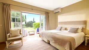 A bedroom with a large bed, a chair, a small table, and a view of the outdoors through a sliding door. - Cegonha Country Club (Loulé)