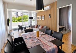 A modern living room with a dining area, large windows, and a view of greenery. - Paraíso - Touristic Apartments (Funchal)