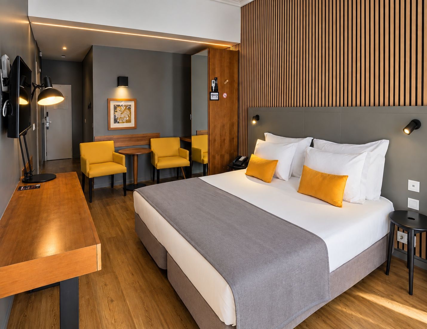Una habitación de hotel con cama, mesitas de noche, un escritorio con silla, una pequeña mesa con sillas y un televisor instalado en la pared.