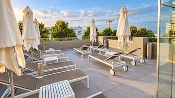 Un área de lounge en la azotea con camas sol y sombrillas, una grúa de construcción al fondo, y un edificio con un letrero.