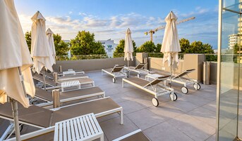 Un área de lounge en la azotea con camas sol y sombrillas, una grúa de construcción al fondo, y un edificio con un letrero.