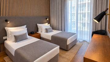 Habitación de hotel con dos camas, un escritorio, una lámpara y vistas a la ciudad a través de amplios ventanales.