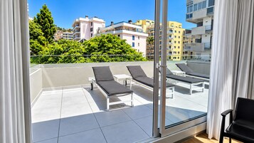 A balcony with sun loungers, a glass door, and a view of buildings and greenery.