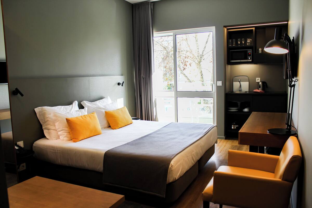 A hotel room with a bed, a desk, a chair, and a window with a view of trees.