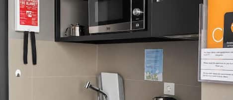 A compact kitchen with a sink, stove, and microwave. There are shelves with dishes and a kettle.
