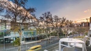 A modern outdoor seating area with white chairs and a glass table, overlooking a street with a yellow taxi and buildings in the background. - Paraíso - Touristic Apartments (Funchal)
