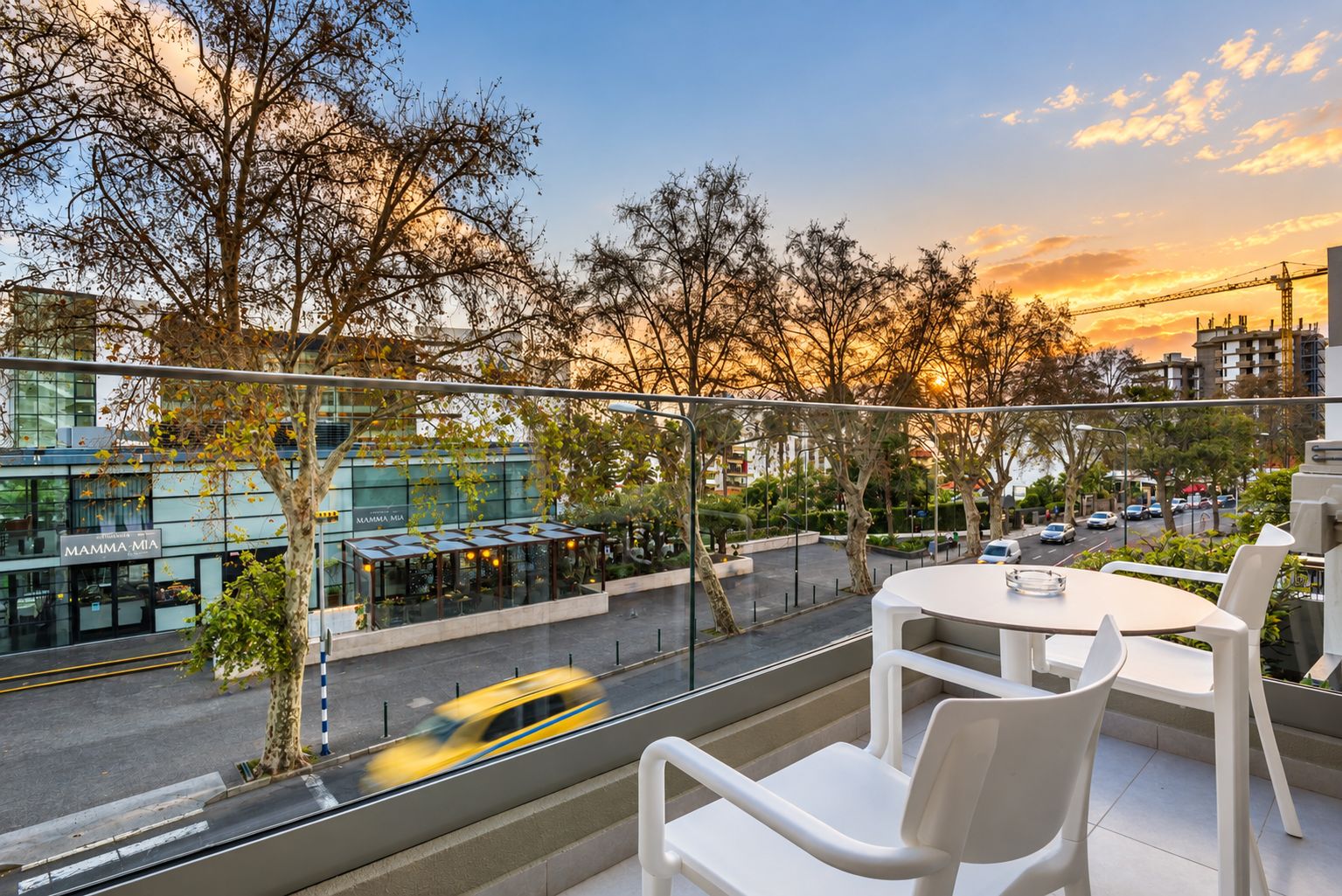 A balcony with white chairs and a table, overlooking a street with a yellow taxi and buildings in the background.