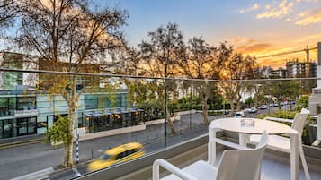 A balcony with white chairs and a table, overlooking a street with a yellow taxi and buildings in the background.
