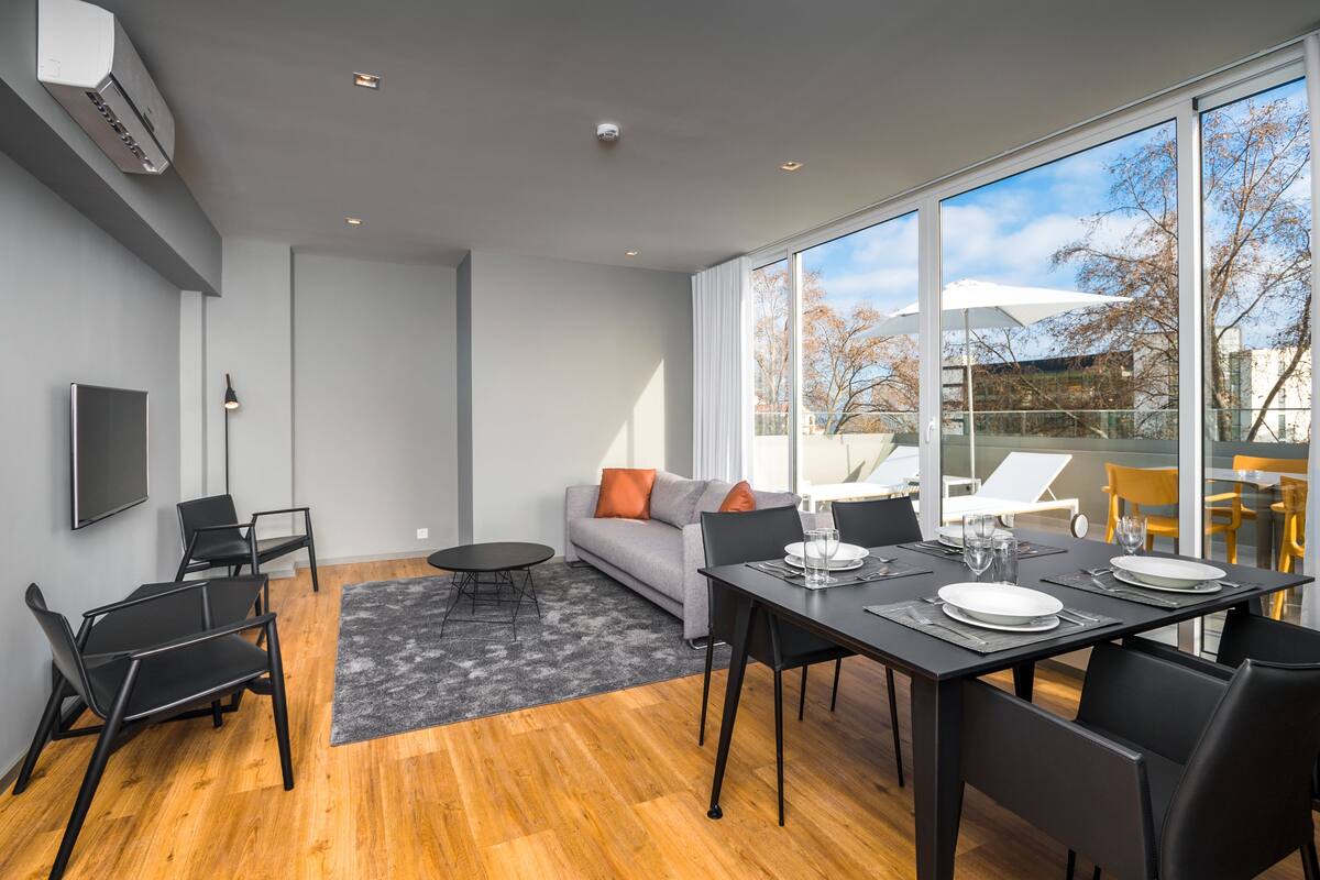 A modern living room with a sofa, dining table, and chairs. There is a TV mounted on the wall, and a large window with a view of the outdoors.