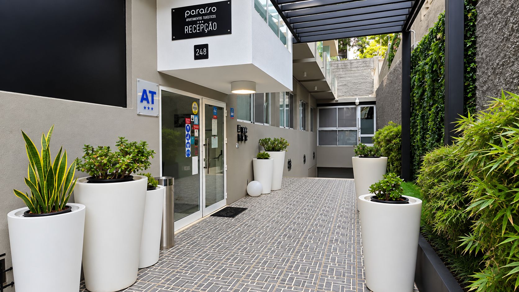 Zona de recepción de hotel con un diseño moderno, que incluye un mostrador de recepción, plantas en macetas y un letrero que indica la ubicación de la recepción.