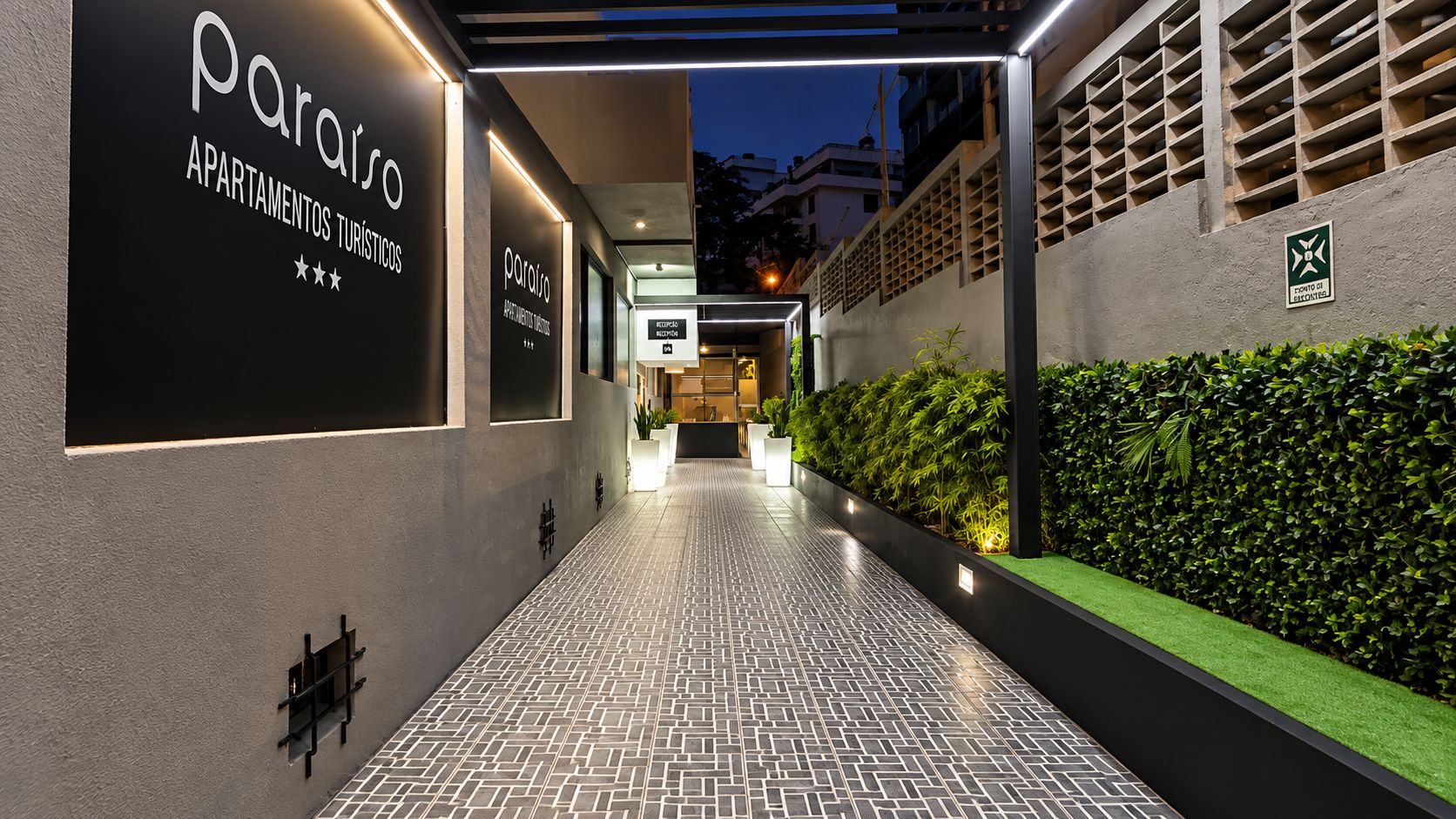 Una entrada de edificio moderno con señalización iluminada para 'Paraiso Apartamentos Turísticos' y un camino que lleva a la entrada.
