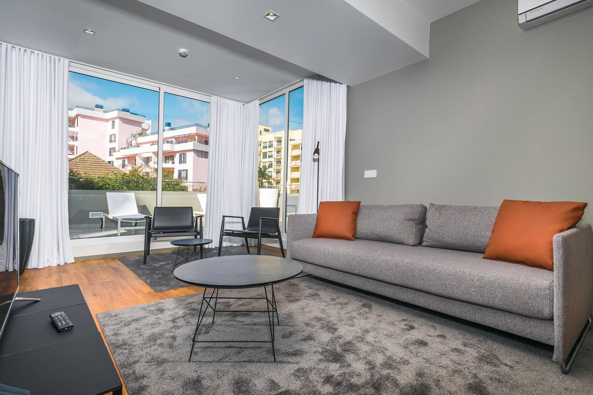 A modern living room with a grey sofa, orange cushions, a round coffee table, and a view of a building through large windows.