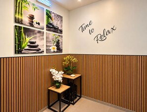 Un área de recepción con un letrero en la pared que dice 'Time to Relax'. Hay cuatro cuadros enmarcados que muestran paisajes naturales tranquilos, y una pequeña mesa de madera con un jarrón de flores blancas.