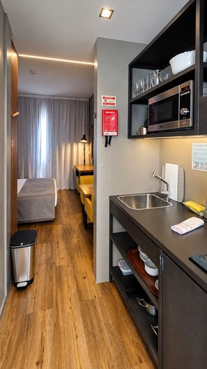 A compact hotel room with a kitchenette, a bed, a bedside table, a trash bin, and a bathroom with a sink and mirror. - Paraíso - Touristic Apartments (Funchal)