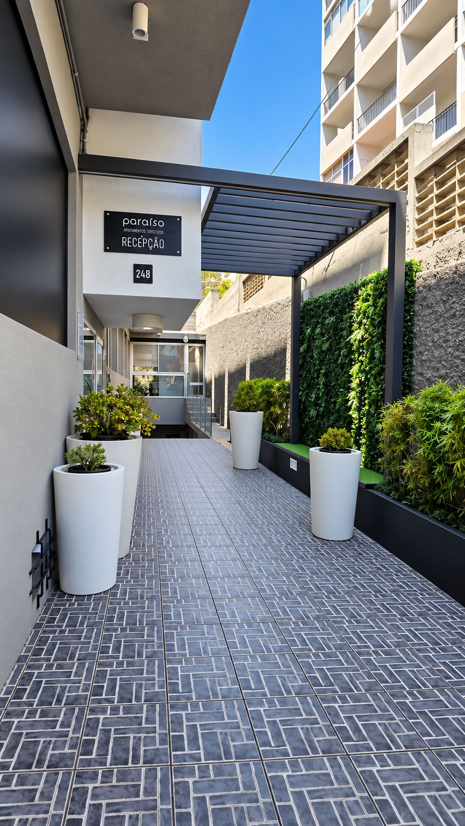 Una entrada moderna a un edificio con letrero de recepción, plantas en macetas y un camino pavimentado con baldosas.