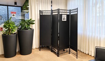 A room with a black partition, two large potted plants, a sign indicating a business center, and a view through a window with a curtain.