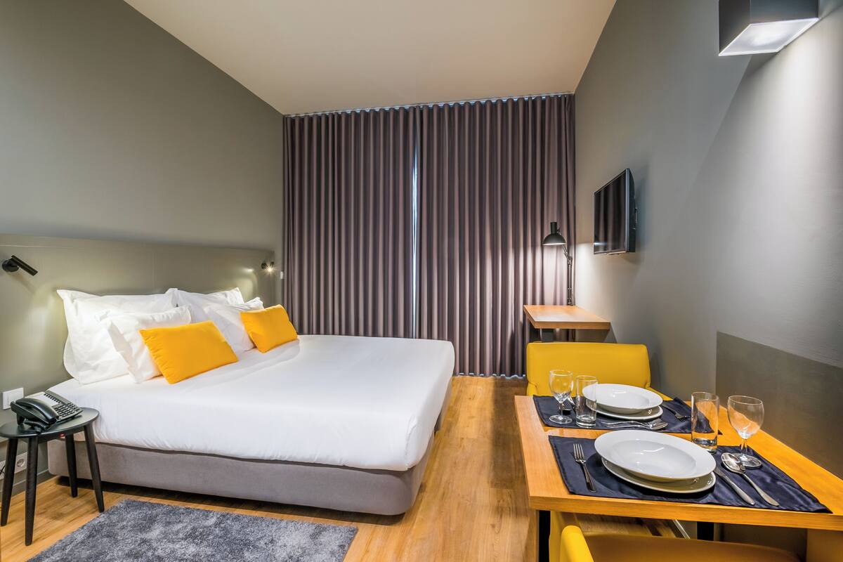 A hotel room with a bed, a desk with a chair, a dining table set with plates and glasses, and a TV mounted on the wall.
