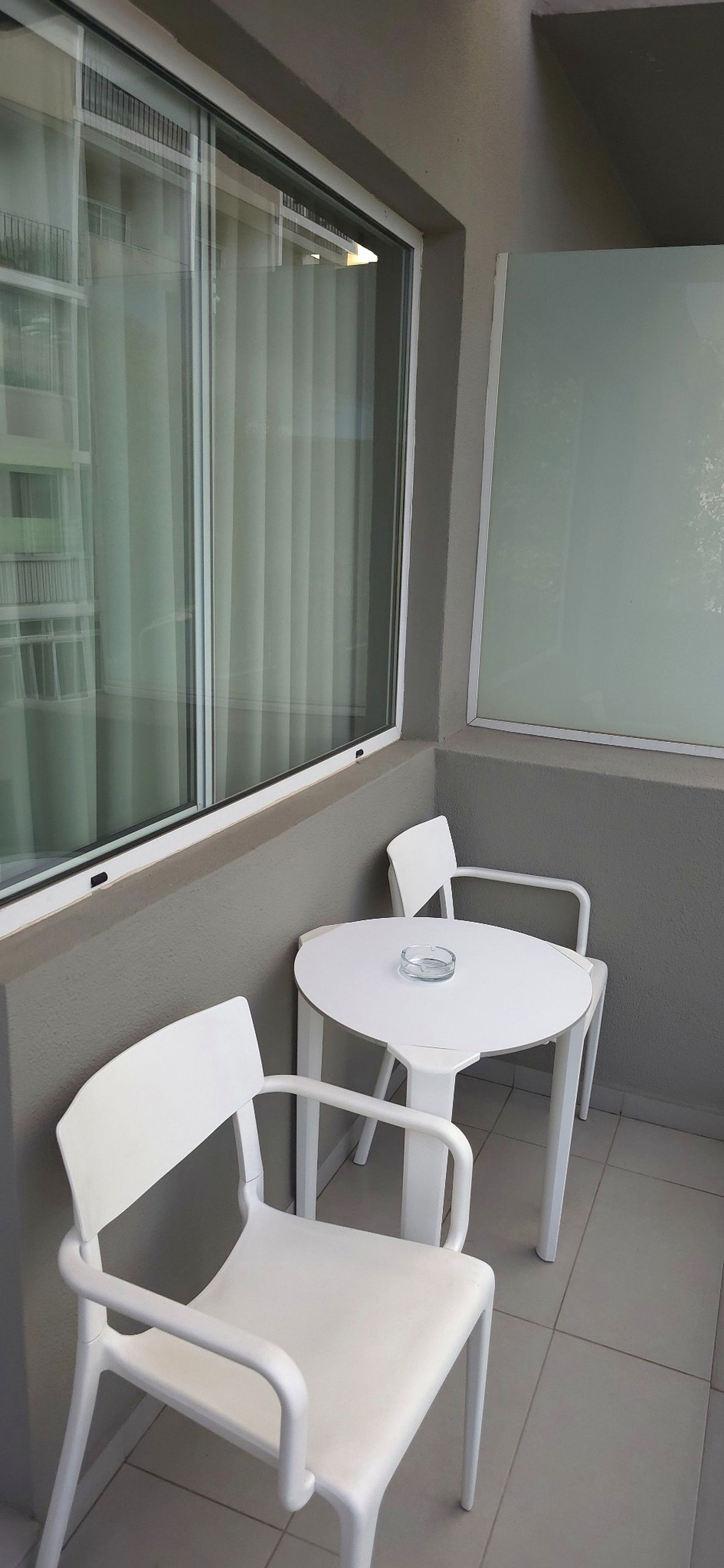 Ein kleiner Balkon mit zwei weißen Stühlen und einem runden Tisch, ein Fenster mit Vorhängen sowie eine Wand mit einer Milchglasscheibe.