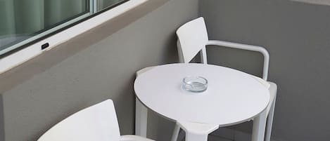 A small balcony with two white chairs and a round table, a window with curtains, and a wall with a frosted glass panel.