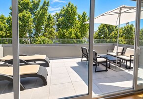 Un balcón moderno al aire libre con asientos, una mesa, puertas de cristal y vistas a árboles.
