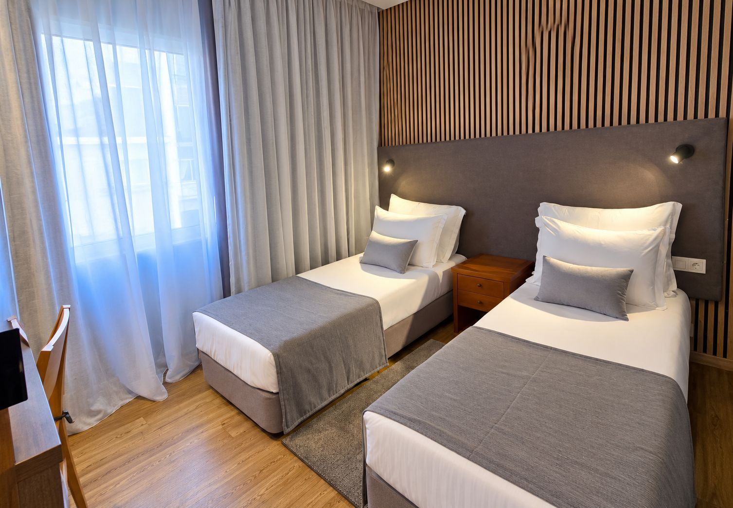 Ein Hotelzimmer mit zwei Betten, Holzboden, einem großen Fenster mit Vorhängen und einer Wand mit vertikalen Holzlatten.