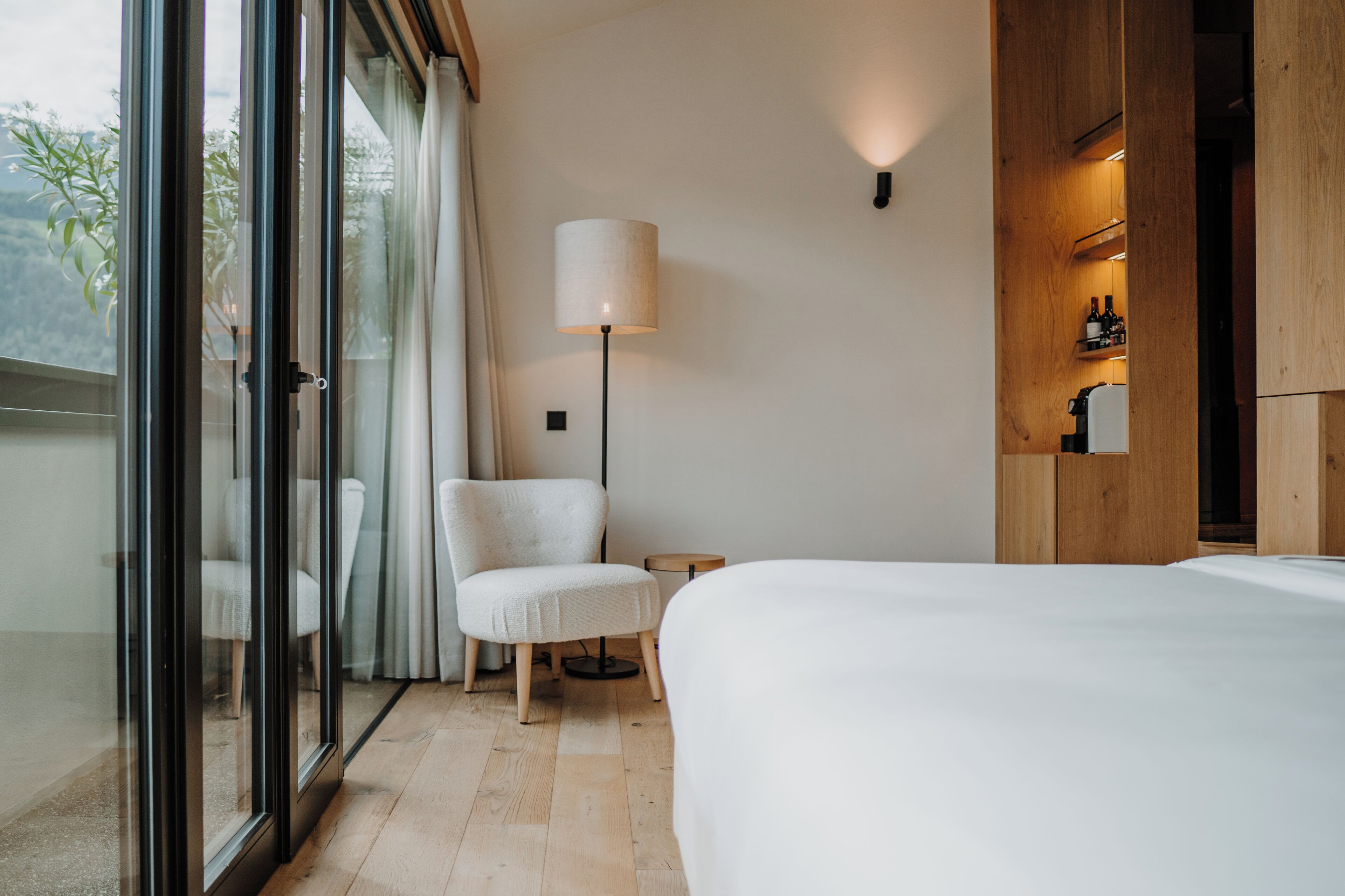 Una habitación de hotel moderna con una cama grande, un sillón blanco, una lámpara de pie y un aparador de madera con botellas.