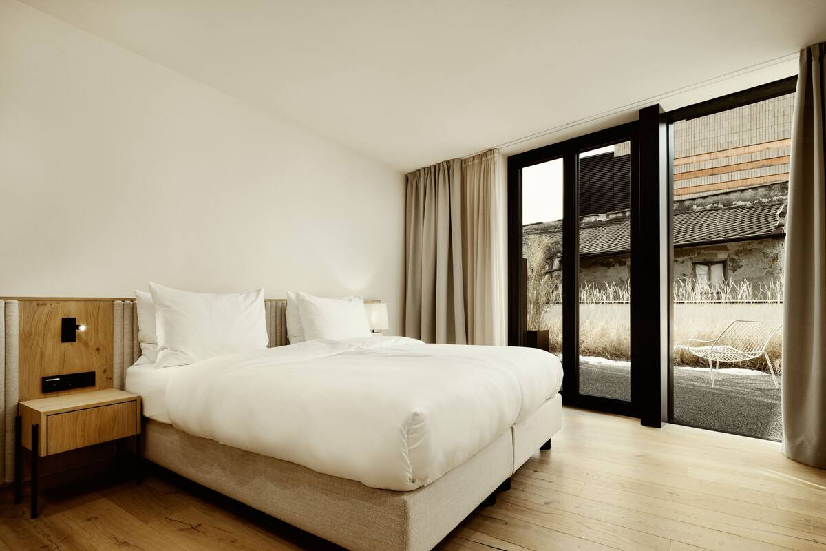 A modern hotel room with a large bed, a bedside table with a lamp, and a view of a traditional building through a glass door.