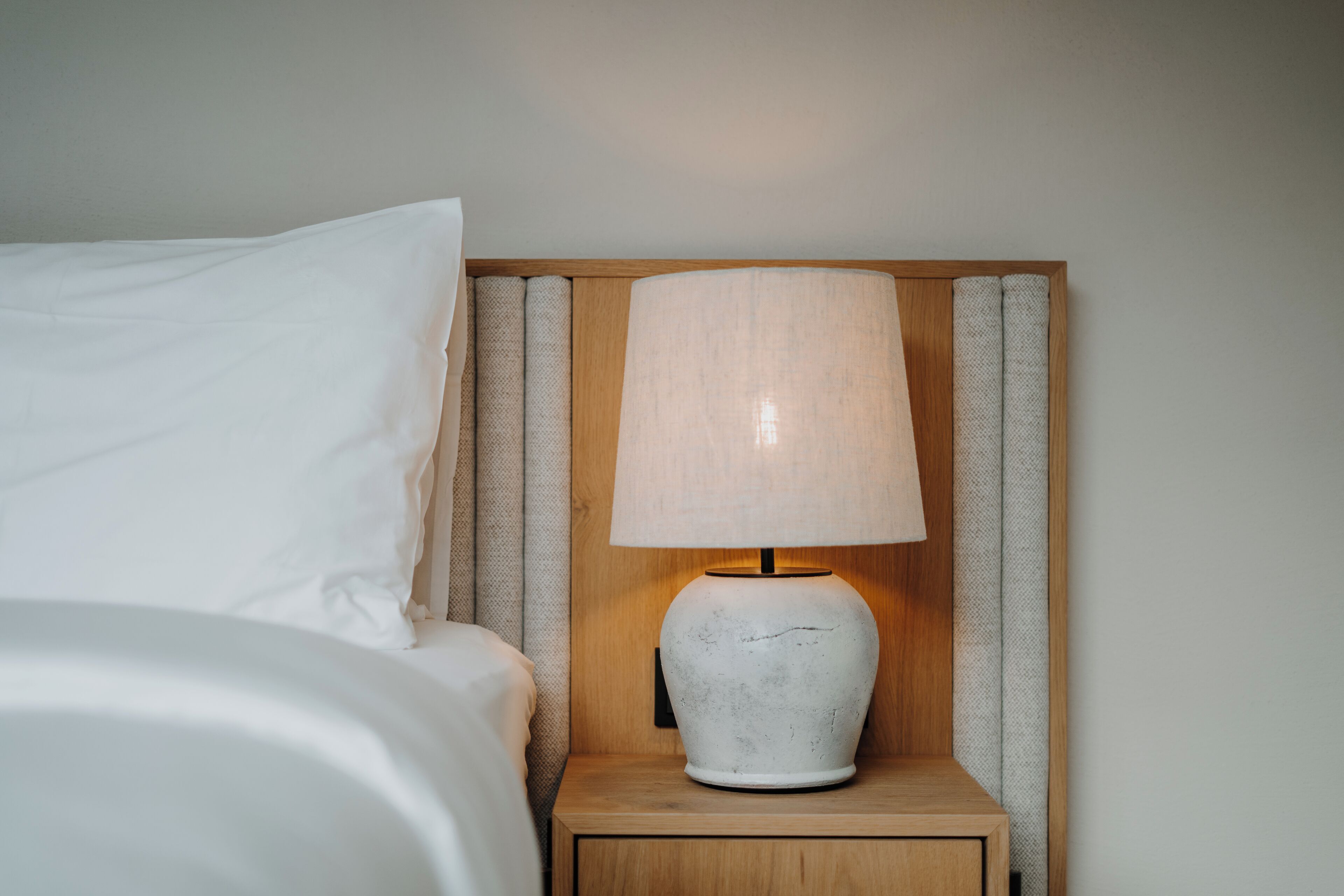 Een netjes opgemaakt bed met witte lakens, een houten nachtkastje met een witte keramieken lamp, en een houten hoofdbord.