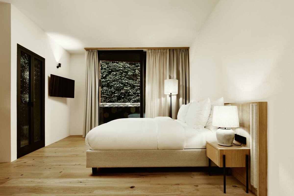 A bedroom with a bed, a nightstand with a lamp, a TV on the wall, and a window with curtains.
