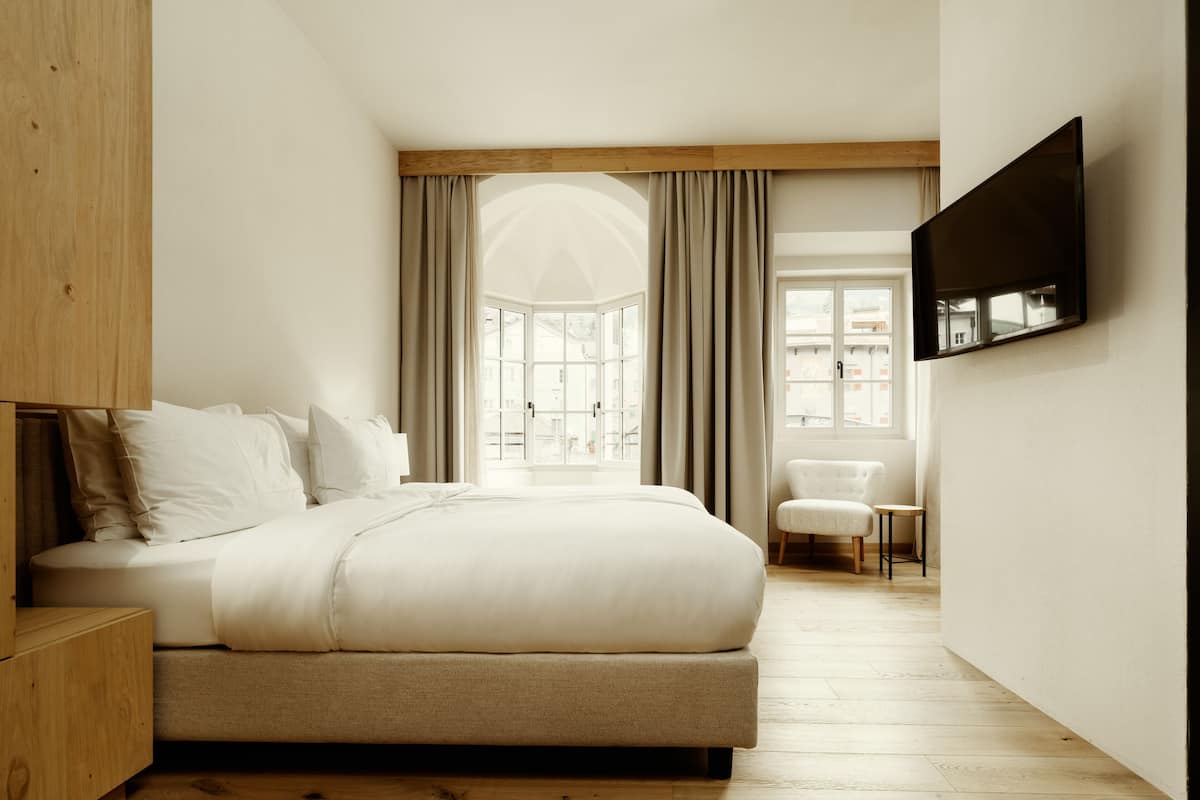 A modern bedroom with a large bed, a wooden nightstand, a chair, a TV mounted on the wall, and a window with curtains.
