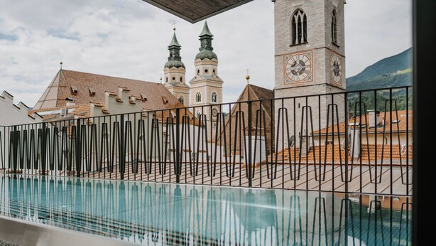 Zona de piscina con una cerca metálica, una torre del reloj y dos torres puntiagudas al fondo.
