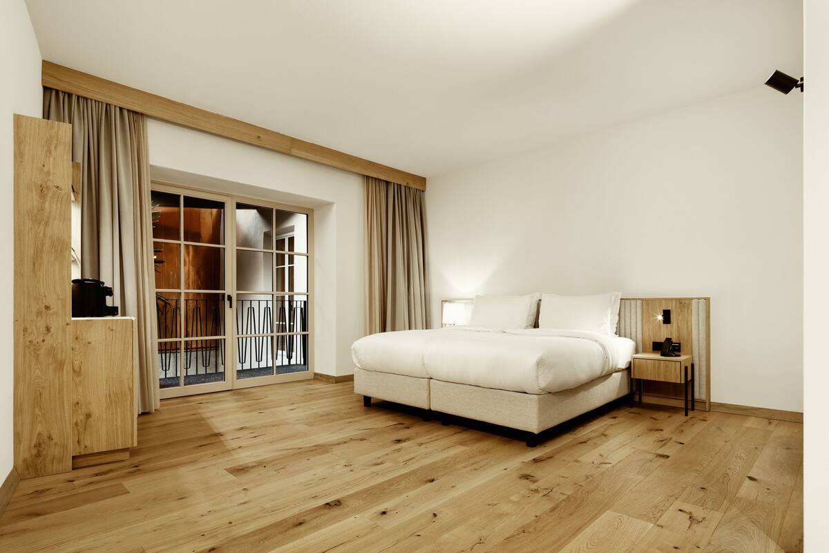 A bedroom with a large bed, wooden flooring, a sliding glass door leading to a balcony, and a small bedside table.